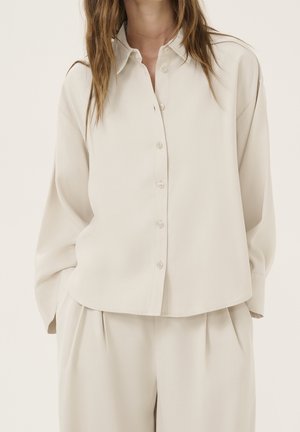Femme portant une chemise beige ample boutonnée et un pantalon large assorti, les mains dans les poches.