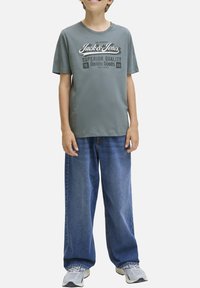 Camiseta de algodón gris con texto y logo en blanco, combinada con unos vaqueros de denim azules sueltos. Ajuste casual con mangas cortas y un escote redondo.