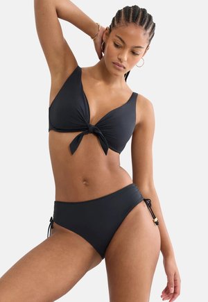 Kvinde med fletninger iført en sort bikini med knude foran på toppen og justerbare bånd i siden på trusserne mod en ensfarvet baggrund.