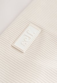 Primo piano di un tessuto a coste color crema con un'etichetta rettangolare bianca al centro, riportante in rilievo la parola "self."