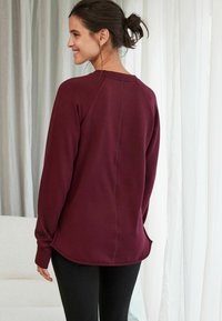 Bordeauxfarbenes Sweatshirt mit entspannter Passform, abgerundetem Saum, Raglanärmeln und subtilen Nähdetails. Weicher Stoff, kombiniert mit schwarzen Leggings.