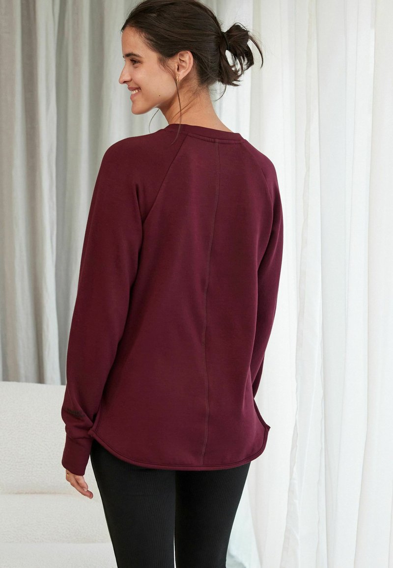 Bordeauxfarbenes Sweatshirt mit entspannter Passform, abgerundetem Saum, Raglanärmeln und subtilen Nähdetails. Weicher Stoff, kombiniert mit schwarzen Leggings.