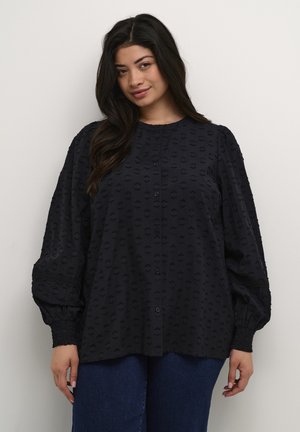 Jūras zila teksturēta blouse ar pogu aizdari priekšā, pārākām piedurknēm un veidotiem aprocēm, ar paaugstinātu punktiņu rakstu visā apģērbā.