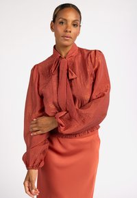 Roodbruine blouse met een striknek, textuurstof, pofmouwen en een geraffineerde zoom, gecombineerd met een bijpassende rok. Gladde, zachte afwerking.
