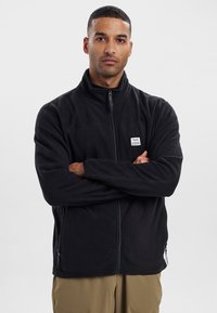 Giacca in pile nera con zip frontale, collo alto, tasche laterali e un piccolo logo ricamato sul lato sinistro. Materiale morbido e testurizzato.
