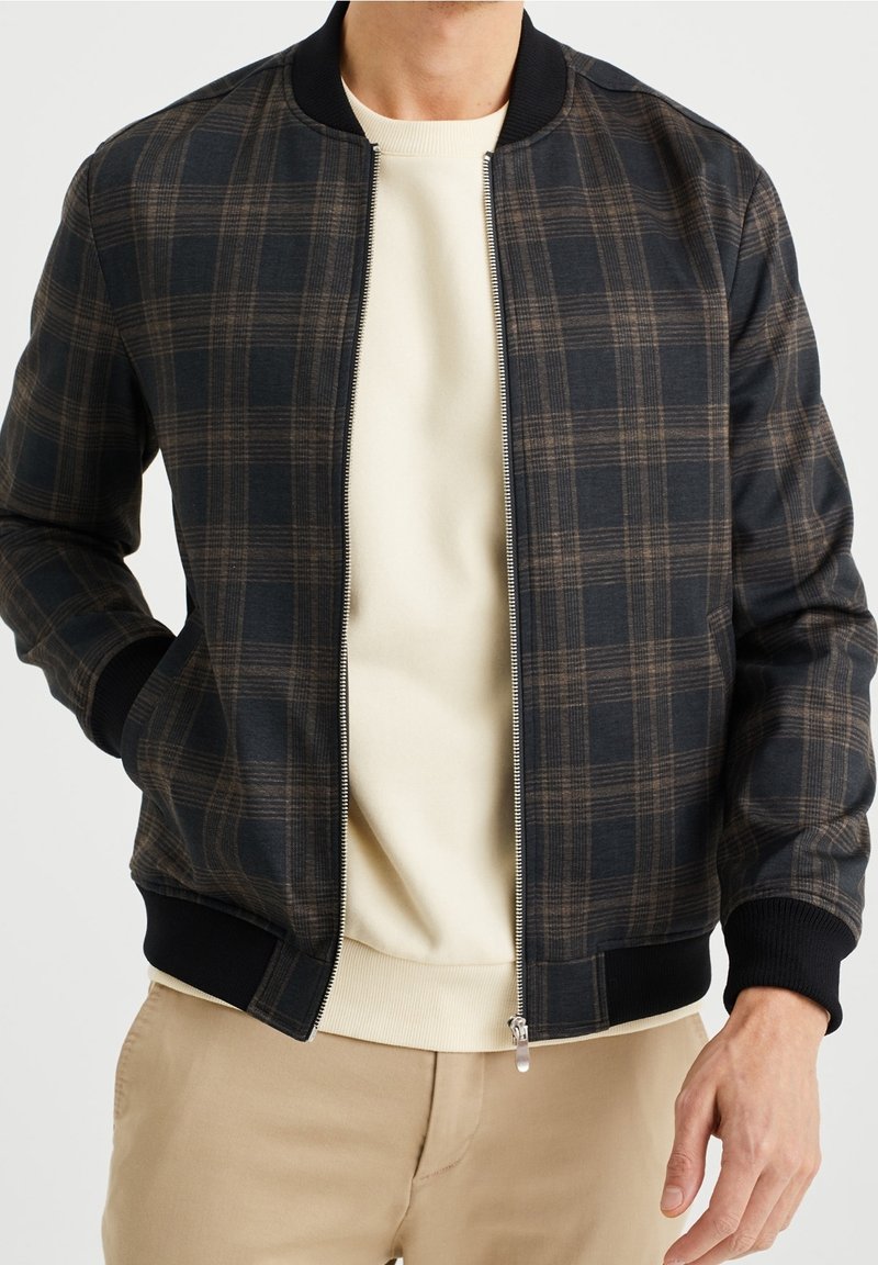 Muž v černo-hnědo-kostkované zipové bundě přes krémovou mikinu a béžových kalhotách, s rukou v kapse, na jednobarevném pozadí.