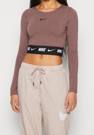 Femme portant un crop top mauve à manches longues avec une bande noire au logo Nike et un pantalon beige à cordon avec un emblème blanc Nike swoosh.