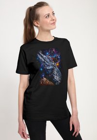 Mladá žena v černém tričku Star Wars s motivem Millenium Falcon, postavami a kosmickým pozadím, stojící před jednoduchou světlou zdí.