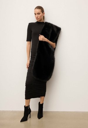 Zwarte bodyconjurk met plooien, gecombineerd met een zwarte bontsjaal die over één schouder is gedrapeerd. Zwarte enkellaarsjes met een puntige neus maken de look compleet.