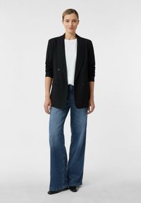 Donna in piedi che indossa un blazer nero, una camicia bianca, pantaloni in jeans larghi blu e scarpe nere a punta, davanti a uno sfondo chiaro e neutro.