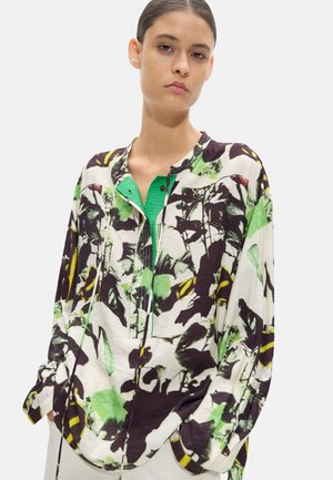 Młoda kobieta ubrana w luźną, długorękawową bluzkę w kwiatowy wzór w kolorach zielonym, czarnym, białym i żółtym, z rękami w kieszeniach.