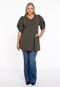 Olivgrünes V-Ausschnitt-Oberteil mit Puffärmeln, aus weichem Stoff. Kombiniert mit hellblauen Schlagjeans und schwarzen spitzen Pumps.
