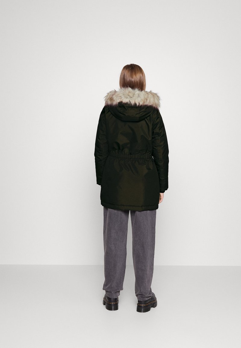 Persona che indossa un parka nero con cappuccio foderato in pelliccia, pantaloni grigi e scarpe nere, in piedi di fronte a una parete bianca semplice.