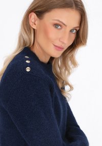 Donna con capelli biondi che indossa un maglione morbido blu navy con tre bottoni dorati decorativi sulla spalla, sorridendo leggermente.