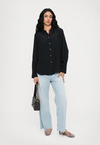 Camisa preta de manga longa com botão, combinada com jeans de perna larga azul claro e sapatos planos listrados multicoloridos. Os acessórios incluem uma bolsa preta.