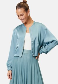 Helle blaue Satin-Bomberjacke mit einem Reißverschluss auf der Vorderseite, weiten Ärmeln, gerippten Bündchen und einem verkürzten Design. Getragen mit einem passenden Rock.