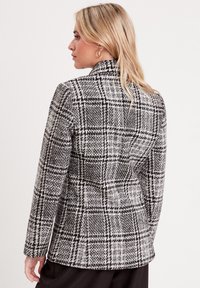 Black and white plaid blazer with a structured collar, long sleeves, and textured fabric. Features a fitted silhouette and tailored design.