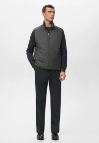 Szary pikowany kamizelka z kołnierzem stojącym, założona na ciemną bluzkę z długim rękawem, w połączeniu z ciemnymi spodniami i czarnymi butami. Gładka faktura, minimalistyczny design.