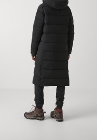 Černý dlouhý péřový kabát s horizontálním prošíváním a kapucí, doplněný o zúžené černé kalhoty a hnědé turistické boty s růžovými akcenty.