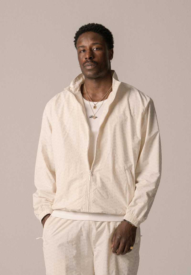 Veste zippée beige clair en tissu texturé, dotée de poignets élastiques et d'un col haut. Portée avec un pantalon assorti et des colliers superposés.