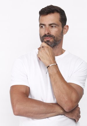 Brățară alb-negru pe încheietura unui braț de culoare maronie, purtată cu un tricou alb simplu. Păr scurt și barbă întunecată. Mână poziționată aproape de bărbie.
