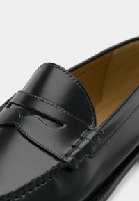 Mustast nahast loafer, millel on ehtsa väärisasjaga detail ja beež sisematerjal, mida iseloomustavad siledad läikivad pinnad ja õmmeldud aktsendid.