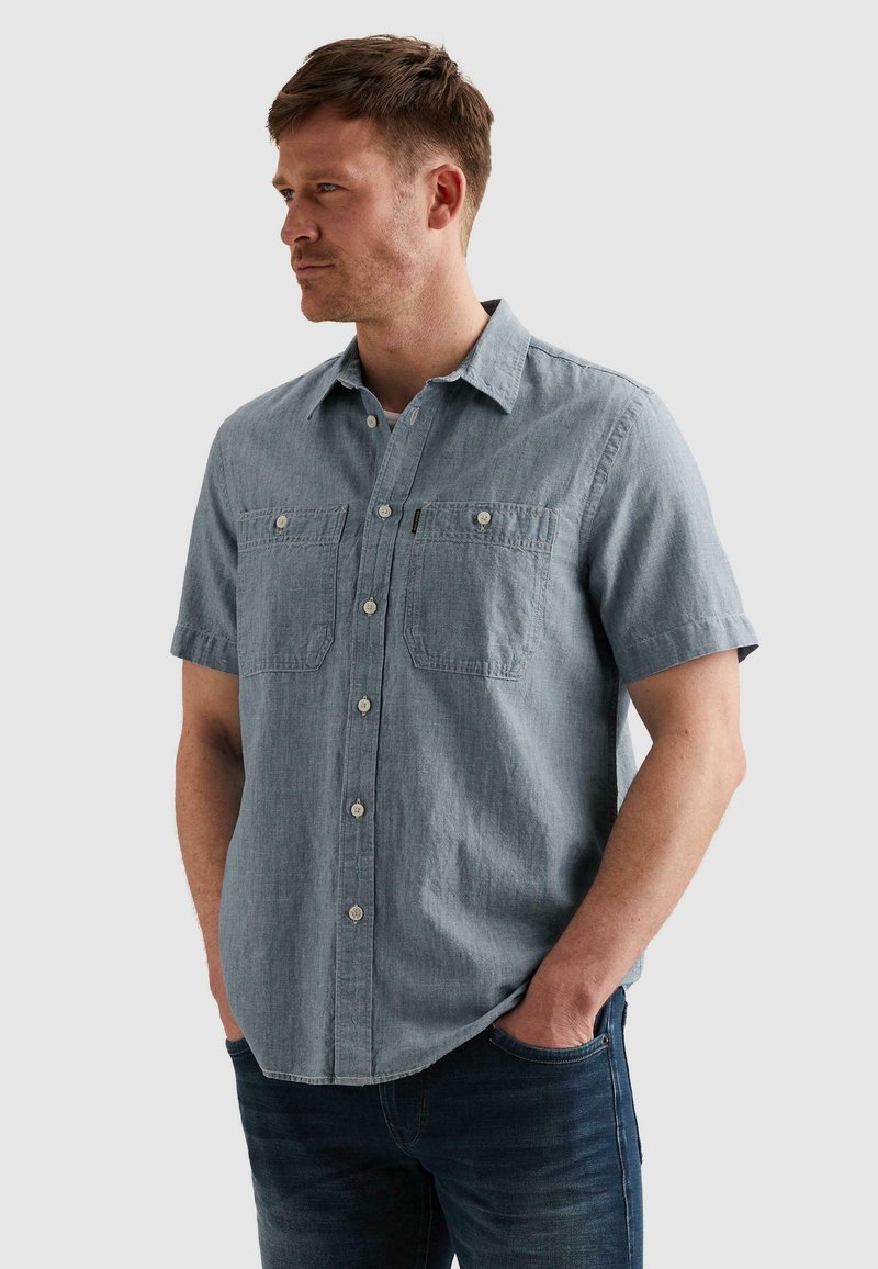 Homme portant une chemise en denim claire à manches courtes avec deux poches poitrine, debout les mains dans les poches d'un jean bleu foncé, regardant vers la gauche.