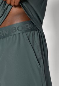 Calções atléticos cinzentos com uma cintura larga onde está impressa a palavra "BORN" num padrão discreto, feitos de um tecido leve e suave, com bolsos laterais.