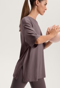 Volné fialové tričko s krátkým rukávem, kulatým výstřihem a bočními rozparky. Modelka drží světle růžovou cvičební kouli, stojí v profilu.