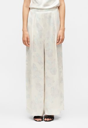 Naine kannab kõrge vöökohaga, laia lõikega elevandiluu värvi pükse, millel on peen lillemuster, ja musta värvi avatud varbaga kontsakingi pitsilise detailiga.