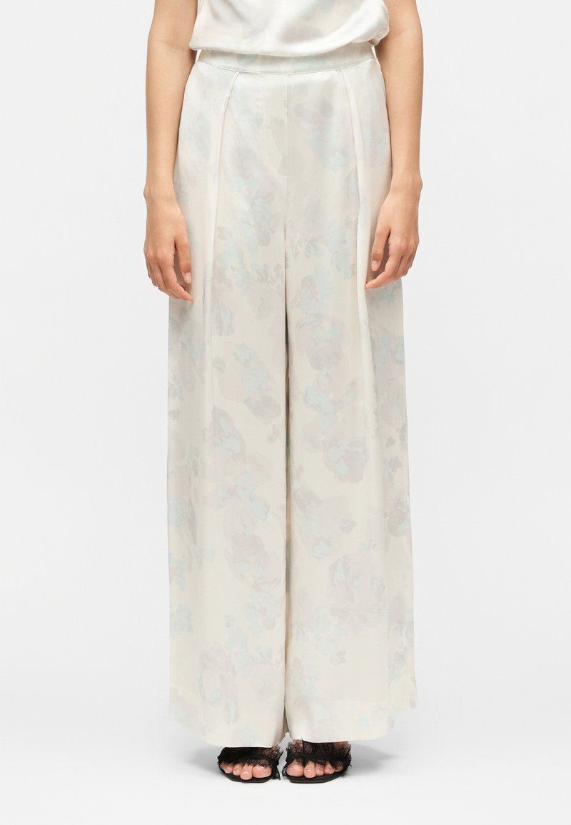 Vrouw draagt een hooggetailleerde, wijde, ivoorkleurige broek met een subtiel bloemenpatroon en zwarte open-teenhakken met kantdetail.