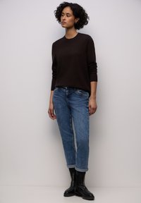 Maglione marrone scuro a maniche lunghe, abbinato a jeans di denim azzurro chiaro con risvolti, e stivaletti neri alla caviglia. Sfondo semplice.