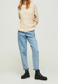 Pull beige en maille avec des motifs géométriques découpés, associé à un jean bleu clair ample et des bottines noires à semelles texturées.