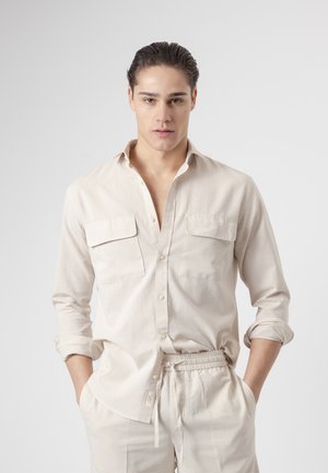 Joven con el pelo peinado hacia atrás lleva una camisa beige clara abotonada con las mangas remangadas y pantalones a juego con cordón, con las manos en los bolsillos.