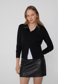 Schwarze Strickjacke mit Knopfverschluss, langen Ärmeln und klassischem Kragen, kombiniert mit einem taillierten schwarzen Lederrock.