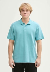 Lichtblauw poloshirt in een textuurstof, voorzien van een kraag, een drieknopenlijst en korte mouwen. G gedragen met donkere broek.