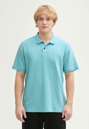 Lichtblauw poloshirt in een textuurstof, voorzien van een kraag, een drieknopenlijst en korte mouwen. G gedragen met donkere broek.
