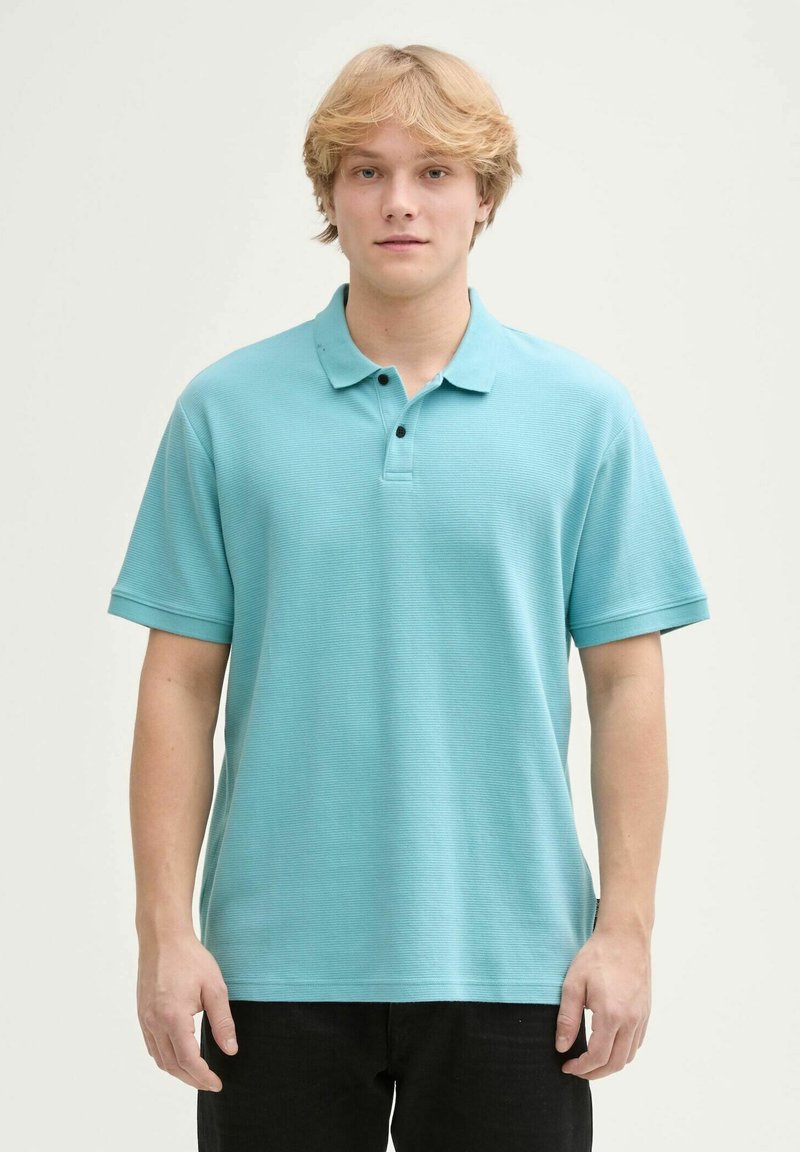 Lysblå polo shirt i et struktureret stof, med krave, tre-knaps placket og korte ærmer. Båret med mørke bukser.