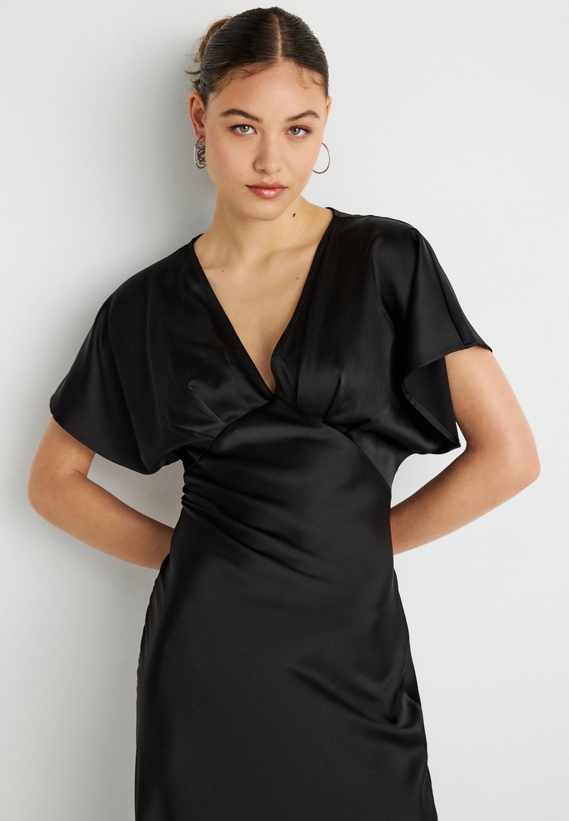Μαύρο σατινέ φόρεμα με βαθύ ντεκολτέ σε σχήμα V, κοντά ανοιχτά μανίκια και ρυτιδωτή λεπτομέρεια στη μέση. Ομαλή υφή, εφαρμοστή σιλουέτα.