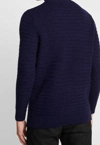 Granatowy sweter dzianinowy z teksturowanym wzorem, ściągaczami na mankietach i dolnej krawędzi. Posiada wysoki kołnierz oraz luźny krój.