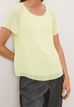 Vrouw draagt een losse, lichtgele blouse met plooien en korte mouwen, gecombineerd met een zwarte broek, staand tegen een effen lichte achtergrond.