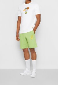 Camiseta blanca con gráfico floral y texto, combinada con pantalones cortos verdes con detalles de logo amarillo. Zapatillas blancas y calcetines completan el conjunto.
