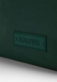 Bolsa de tela de color verde oscuro con un parche de cuero texturizado con "SCALPERS" en letras mayúsculas. Superficie lisa, diseño minimalista.