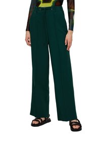 Person wearing dark green wide-leg pants with front seams and black open-toe sandals, standing with hands relaxed on sides.