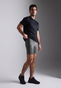 Menns svart treningst-skjorte kombinert med grå tettsittende shorts. Stoffet ser glatt ut med minimale sømmer. Brukt med svarte joggesko.