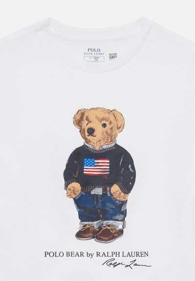 Balta T-kreklu ar multfilmu lāci melnā džemperī ar Amerikas karogu, zilām biksēm un brūnām kurpēm. Iekļauts Ralph Lauren logo.