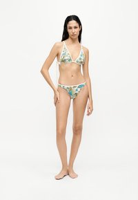 Femme aux longs cheveux foncés portant un bikini blanc avec un imprimé floral bleu et vert, debout pieds nus devant un fond clair uni.