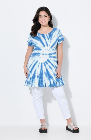 Túnica de vestido en azul y blanco con efecto tie-dye, con un dobladillo acampanado, mangas cortas, combinada con pantalones blancos y sandalias plateadas. Textura de tela suave.