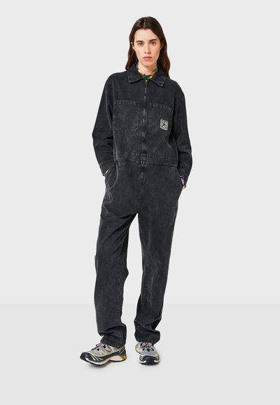 Schwarzer Denim-Overall mit Reißverschluss vorne, zwei Brusttaschen und einem Logopatch. Hat eine lockere Passform und eine strukturierte Oberfläche. Kombiniert mit mehrfarbigen Sneakers.