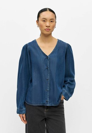 Femme aux cheveux foncés en queue de cheval basse, portant un chemisier en jean bleu à boutons avec des manches longues bouffantes et un pantalon noir, debout sur fond blanc.